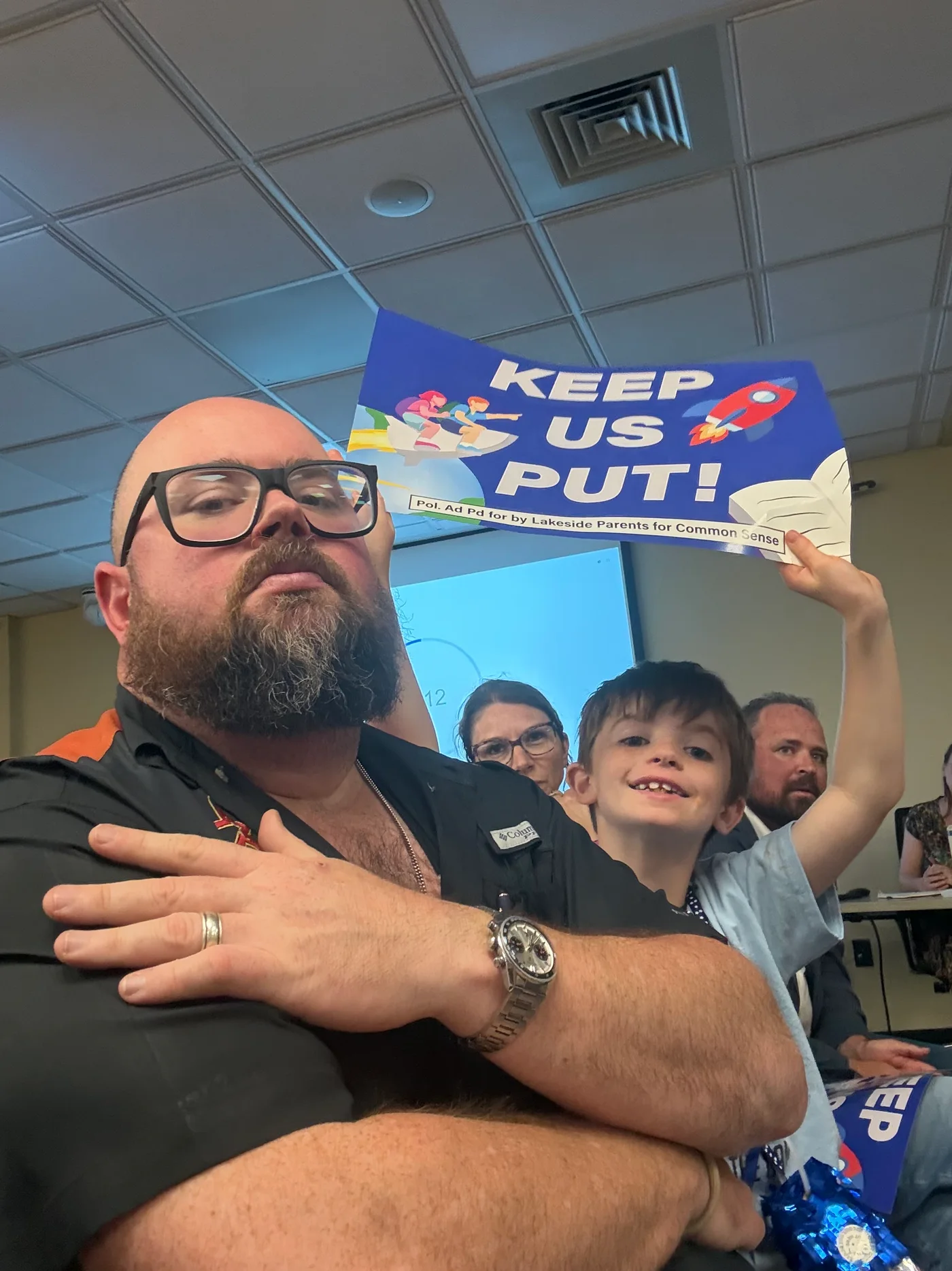 A parent and child at a district meeting, holding a blue Keep Us Put sign.
